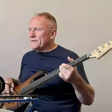 Man in a dark t-shirt plays a bass guitar beside a microphone in a home studio setup.