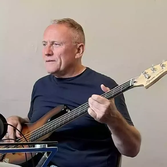 Man in a dark t-shirt plays a bass guitar beside a microphone in a home studio setup.